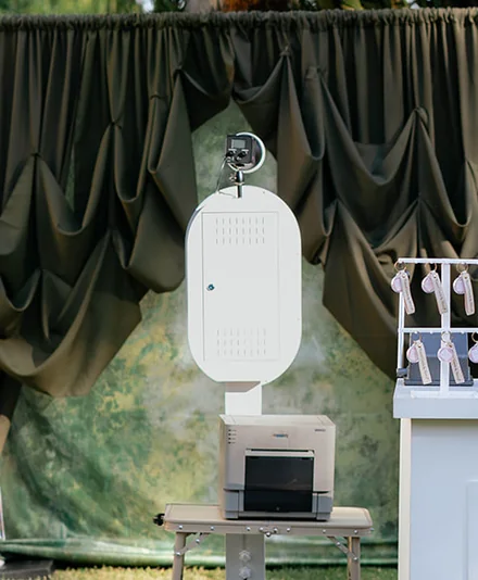Open air photo booth and printer set up outdoors against a draped backdrop.