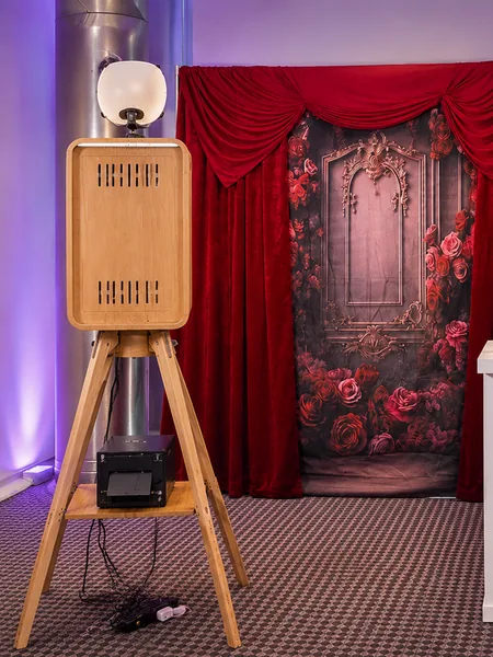 Photo booth on a wooden tripod beside a red velvet and rose backdrop.