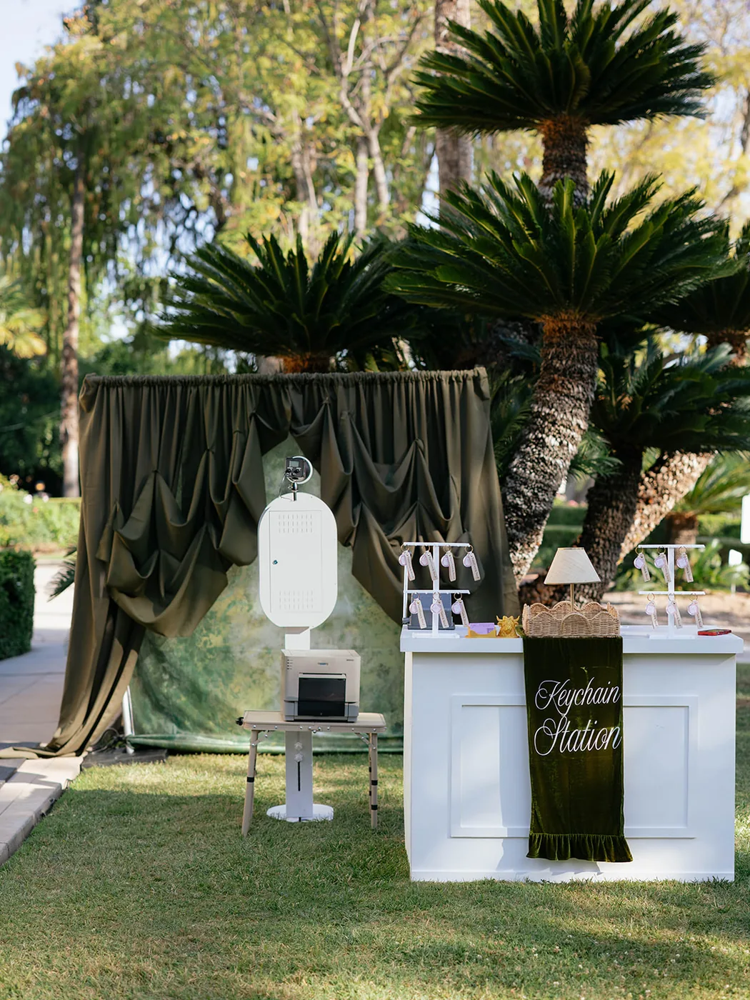 Open air photo booth set up outdoors under palms against a draped backdrop.