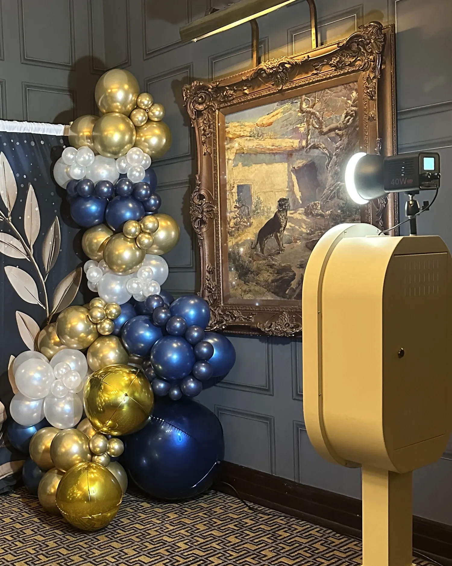 The open air photo booth standing with its light on beside a navy and gold balloon garland in a panelled room.
