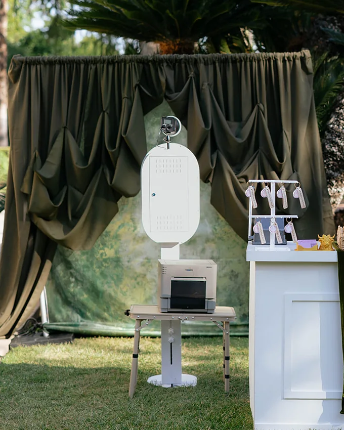 The open air booth outdoors on the grass, with its printer, in front of a draped olive backdrop.