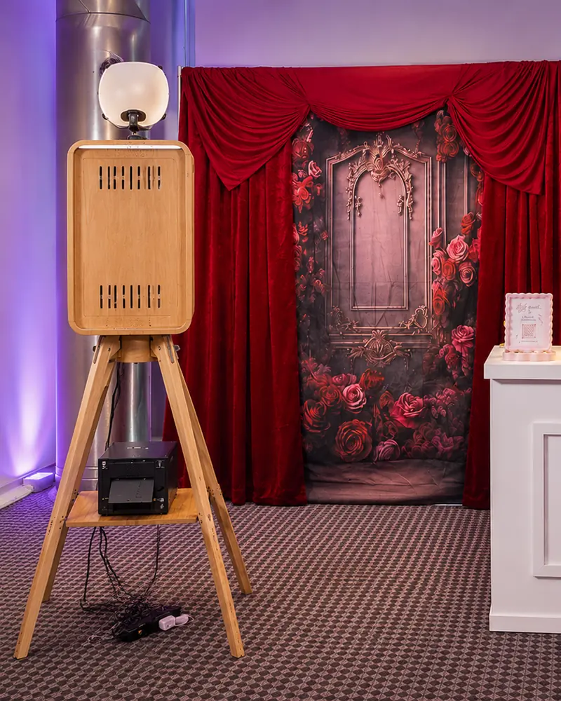 The open air booth and its printer in front of a red velvet and rose backdrop.