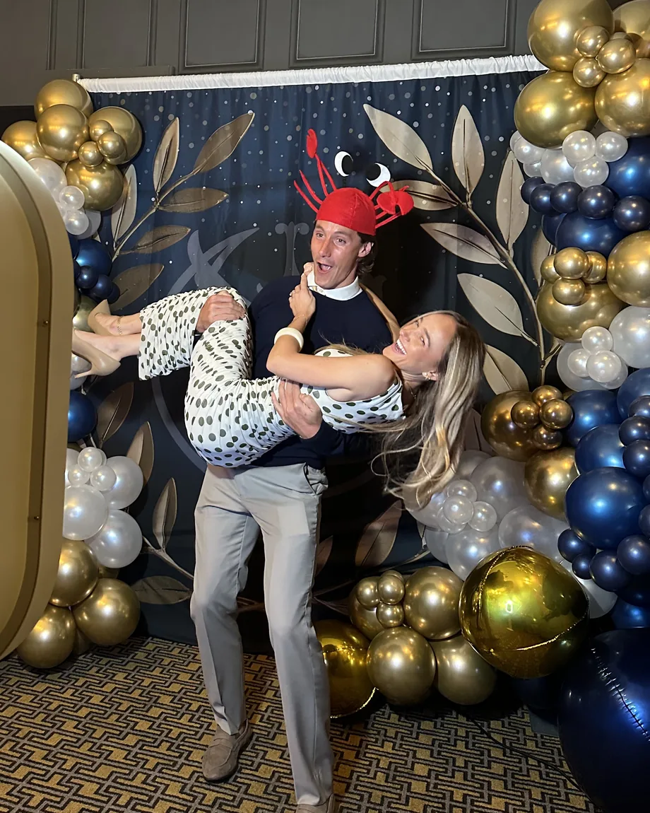 Two guests laughing together in front of a navy and gold balloon backdrop beside the open air photo booth.