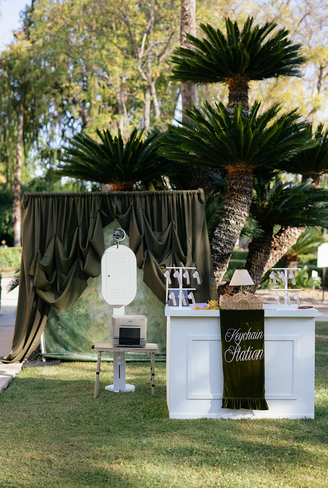 The open air photo booth, printer and keychain bar set up under palms on a lawn.